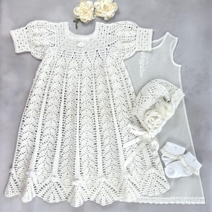 May include: A white crochet dress with short sleeves and a scalloped hem, accompanied by a matching bonnet with a satin ribbon tie and floral embellishments, a pair of white socks with lace trim, and a plain white slip dress underneath.
