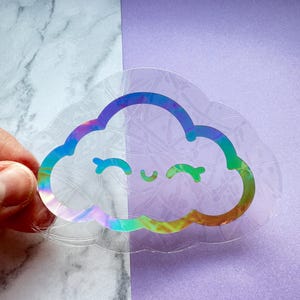 May include: A holographic iridescent sticker of a cloud with a smiling face. The sticker is clear and has a textured background.