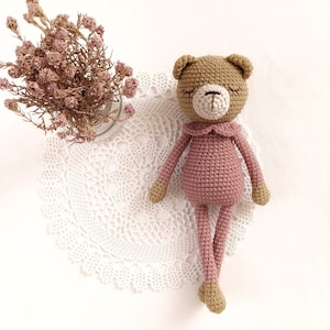 May include: A crocheted pink bear with brown eyes and a brown nose. The bear is wearing a pink crocheted dress with a white crocheted collar. The bear is sitting on a white crocheted doily.