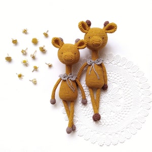 Könnte beinhalten: Zwei gehäkelte Giraffenspielzeuge mit braunen und weißen Akzenten. Die Giraffen sitzen auf einer weißen Häkeldeckchen.