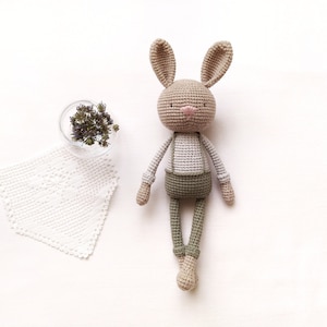 Puede incluir: Un juguete de conejo de crochet con pelaje marrón, una camisa blanca y pantalones verdes. El conejo está acostado de espaldas con los brazos y las piernas extendidos.