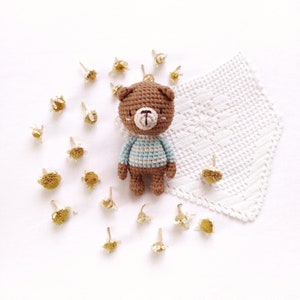 May include: A brown crocheted teddy bear wearing a blue and white striped shirt. The bear is sitting on a white crocheted doily surrounded by dried flowers.
