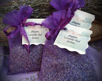 May include: Two purple organza sachets filled with dried lavender buds, each tied with a matching purple ribbon. A white rectangular tag is attached to each sachet, featuring a printed illustration of a red barn and the text Organic Lavender Bud Sachet.