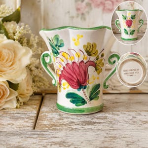 May include: A white ceramic vase with a scalloped rim and two side handles, featuring a hand-painted red flower with green leaves and yellow accents. The base is marked with the text HAND PAINTED IN ITALY FOR FTD in black lettering.