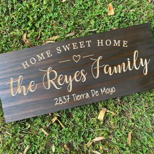 May include: A dark brown wooden sign with the text "Home Sweet Home" and a small heart symbol. Below the text is the name "the Reyes Family" and the address "2837 Tierra De Moyo".