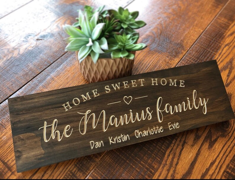 May include: A dark brown wooden sign with the text "Home Sweet Home" and a heart symbol. Below the text is "the Manius family" with the names "Dan, Kristin, Charlotte, Evie" listed underneath.