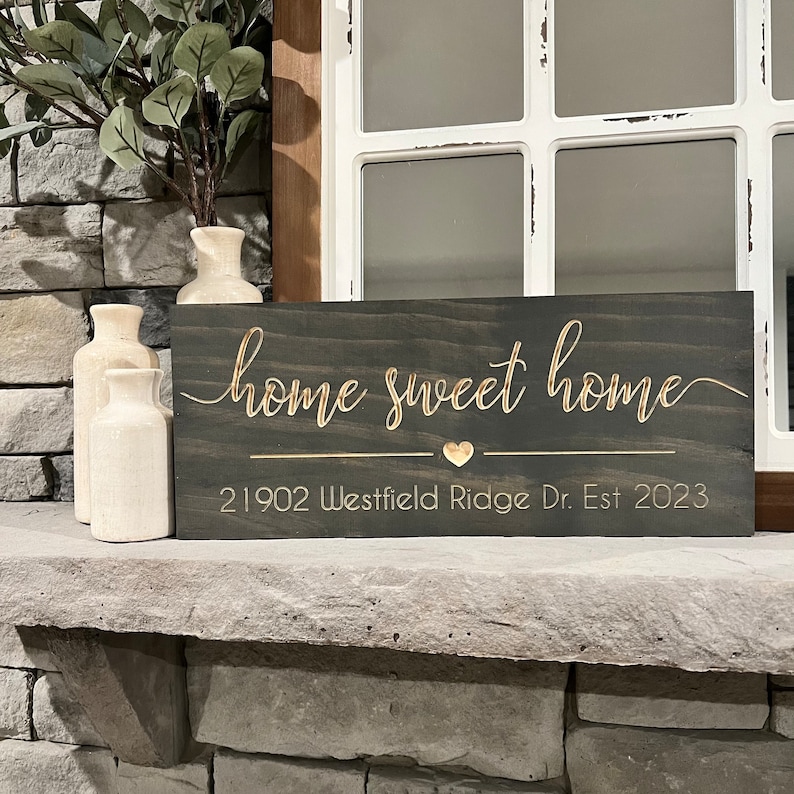 以下が含まれることがあります： 「home sweet home」という金色の文字が書かれた濃い茶色の木製看板。看板には小さな金色のハートと「21902 Westfield Ridge Dr. Est 2023」という金色の文字も書かれている。
