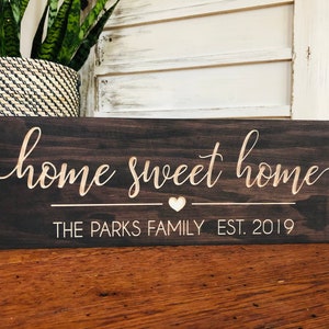 以下が含まれることがあります： 「home sweet home」という金色の文字が書かれた濃い茶色の木製看板。文字の下には小さなハートと「THE PARKS FAMILY EST. 2019」という文字があります。