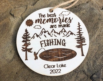 May include: White wooden ornament with a brown wood-burned design of a lake, mountains, trees, a sun, clouds, a fisherman, and the text "The best memories are made fishing Clear Lake 2022".