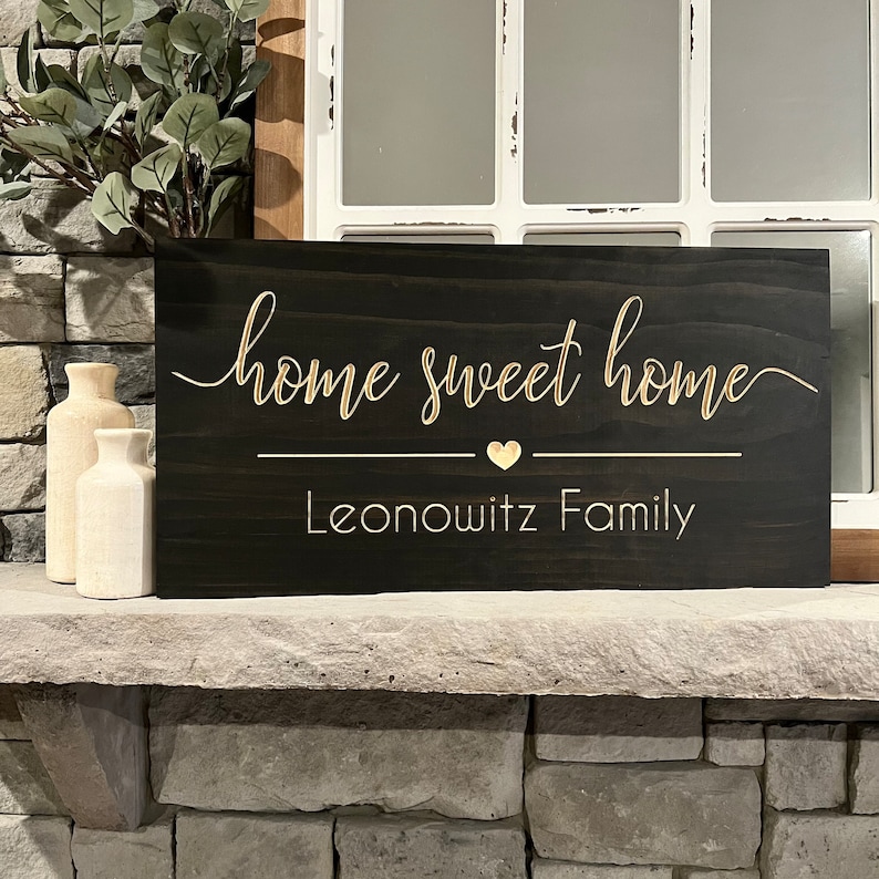 以下が含まれることがあります： 「home sweet home」という金色の文字が書かれた黒い木製の看板。テキストの下には小さな金色のハートと「Leonowitz Family」という金色の文字があります。