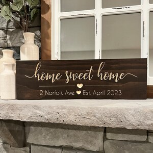 以下が含まれることがあります： 「home sweet home」という金色の文字が書かれた濃い茶色の木製看板。テキストの下には「2 Norfolk Ave」と「Est. April 2023」と、2つの金色のハートが書かれています。