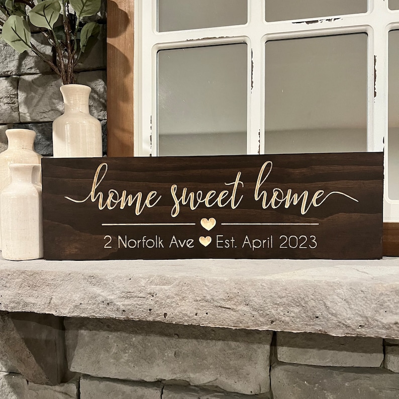 以下が含まれることがあります： 「home sweet home」という金色の文字が書かれた濃い茶色の木製看板。テキストの下には「2 Norfolk Ave」と「Est. April 2023」と、2つの金色のハートが書かれています。