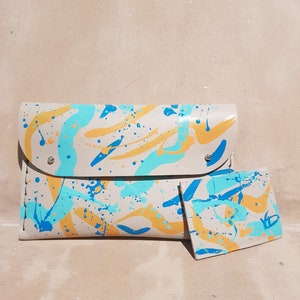 May include: A beige leather clutch with a matching card holder. Both are decorated with a splatter paint design in blue, yellow, and white.