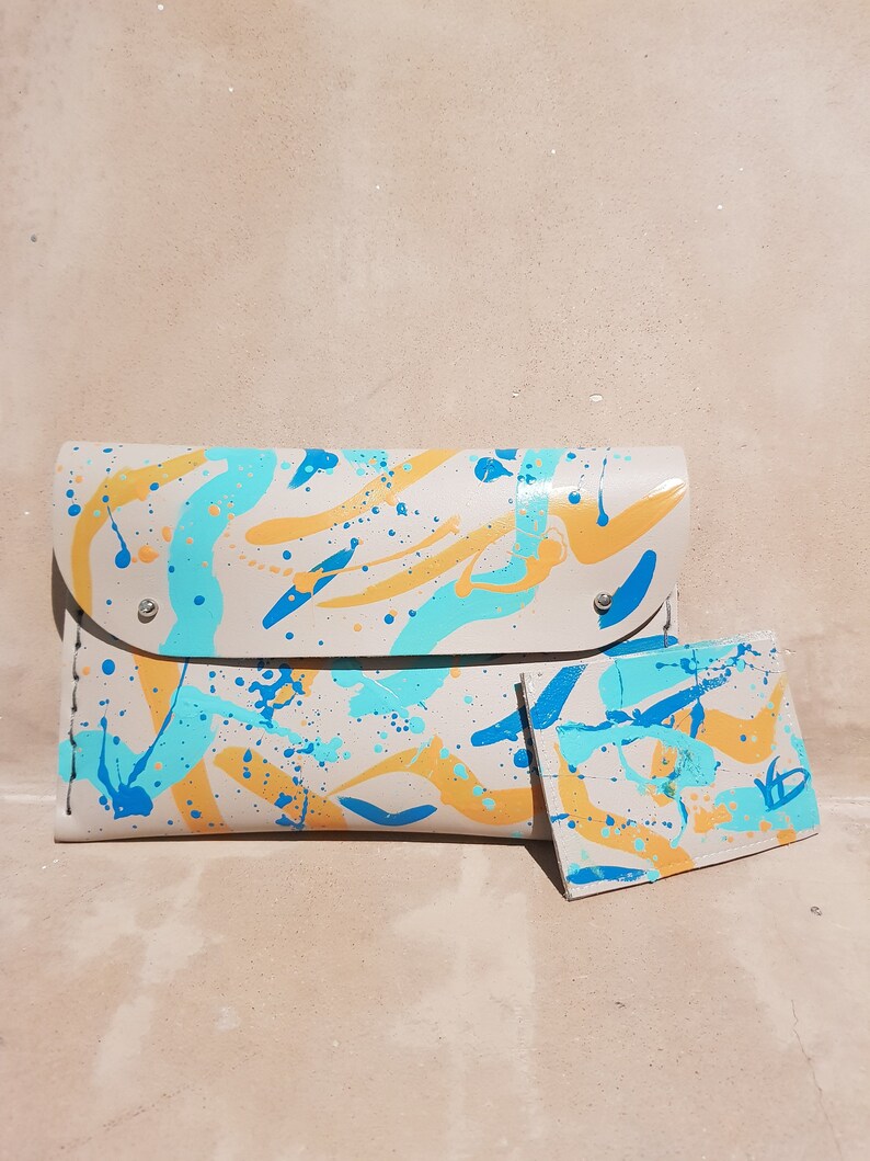 May include: A beige leather clutch with a matching card holder. Both are decorated with a splatter paint design in blue, yellow, and white.