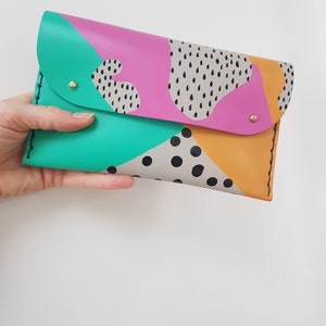 May include: A colorful leather clutch with a geometric pattern in shades of pink, green, yellow, and black. The clutch has a flap closure and gold-toned hardware.