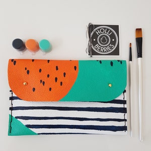 May include: A DIY leather clutch with a watermelon design. The clutch is orange, green, and white with black stripes. It comes with three small paint pots and two paintbrushes. The label says "Holli Berries".