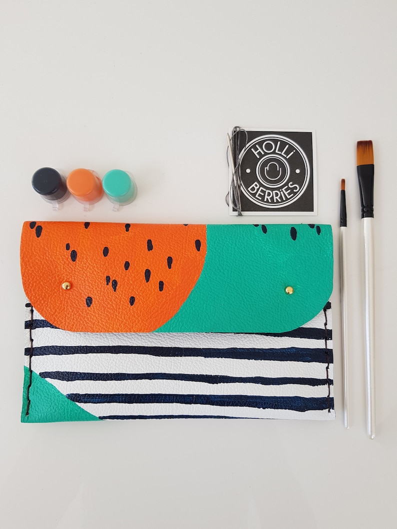 May include: A DIY leather clutch with a watermelon design. The clutch is orange, green, and white with black stripes. It comes with three small paint pots and two paintbrushes. The label says "Holli Berries".
