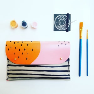 May include: A striped fabric clutch with a pink and orange geometric design. The clutch has a gold zipper and a gold button closure. The clutch is surrounded by paint supplies, including paint tubes and paintbrushes.