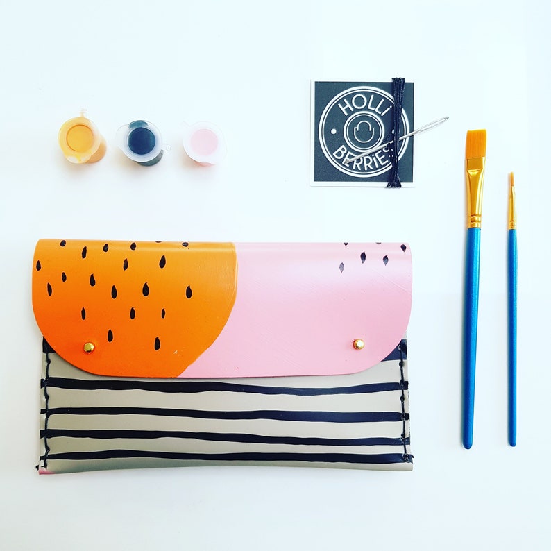 May include: A striped fabric clutch with a pink and orange geometric design. The clutch has a gold zipper and a gold button closure. The clutch is surrounded by paint supplies, including paint tubes and paintbrushes.