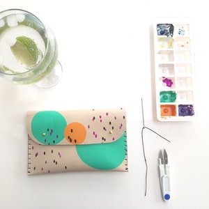 May include: A beige leather wallet with a colorful abstract design featuring circles and dots in shades of green, orange, and black. The wallet is lying on a white surface with a needle, thread, and small scissors.