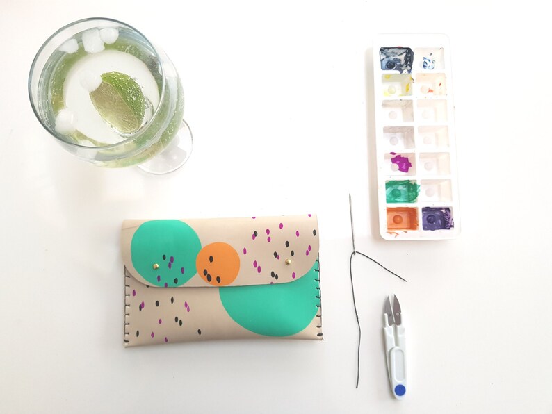 May include: A beige leather wallet with a colorful abstract design featuring circles and dots in shades of green, orange, and black. The wallet is lying on a white surface with a needle, thread, and small scissors.