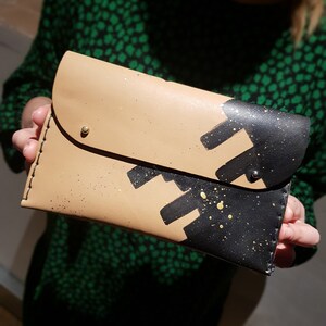May include: A tan leather clutch with black and gold paint splatter design. The clutch has a flap closure and a gold metal button.