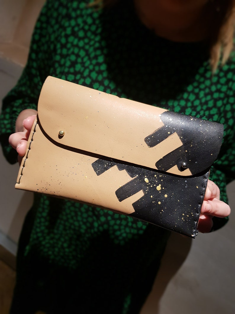 May include: A tan leather clutch with black and gold paint splatter design. The clutch has a flap closure and a gold metal button.