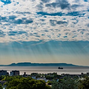 May include: A panoramic view of a city skyline with a large body of water in the foreground. The water is calm and reflects the blue sky and white clouds. A small ship is visible in the distance. The city skyline is in the background and is made up of buildings and trees. The sky is a bright blue with white clouds and sunbeams shining through.