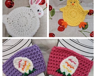 Op de afbeelding: Een verzameling handgemaakte gehaakte items met Paasthema's. Inclusief een witte konijn, een gele kuiken en twee bekerhoezen in paars en roze, elk met een geborduurd eierontwerp. De items worden gepresenteerd op een stof met kersenpatroon.