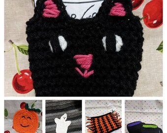 May include: A collection of handmade crochet items, including a black cat face, an orange pumpkin with a smiling face, a white ghost, an orange and black striped coaster, and two black mugs with purple and green accents.