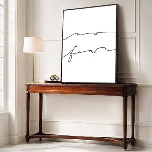 Peut inclure: Une impression d'art encadrée avec les mots "no fear" en écriture noire sur fond blanc. L'œuvre est exposée sur une console en bois foncé avec une lampe et des objets décoratifs.