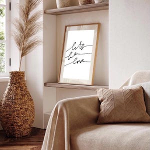 Peut inclure: Une impression encadrée blanche avec le texte "Let's be love" en écriture cursive noire. L'impression est accrochée à une étagère marron clair dans une pièce avec un canapé beige et un vase haut avec de l'herbe de pampa séchée.