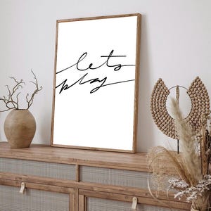Peut inclure: Impression sur cadre blanc avec l'inscription "let's play" en écriture cursive noire. L'œuvre est exposée sur un meuble en bois avec une façade tissée, à côté d'objets décoratifs tels qu'un vase et une décoration murale.