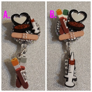May include: Two badge reels with a medical theme. The first reel features a heart-shaped mirror, a red and black makeup compact, a bandage, and a syringe. The second reel features a heart-shaped mirror, a red and black makeup compact, a bandage, and two blood vials. Both reels are decorated with rhinestones.