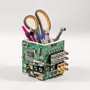 May include: A square desk organizer constructed from green circuit boards featuring various electronic components, including VGA ports, RCA jacks, and integrated circuits. The container holds a pair of scissors with yellow handles, several pens, and a metal tool.