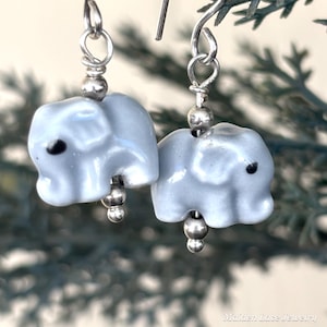 May include: A pair of light blue elephant earrings with black eyes and silver-colored accents. The earrings are suspended from silver-colored hooks and feature small, round beads. The earrings are a whimsical accessory.