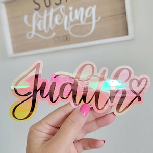 May include: A holographic iridescent pink and gold vinyl sticker with the name "Judith" and a small heart.
