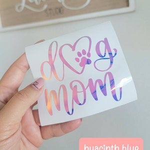May include: A white vinyl sticker with a pink and blue iridescent finish. The sticker says "Dog Mom" with a pink heart and a paw print inside the heart.