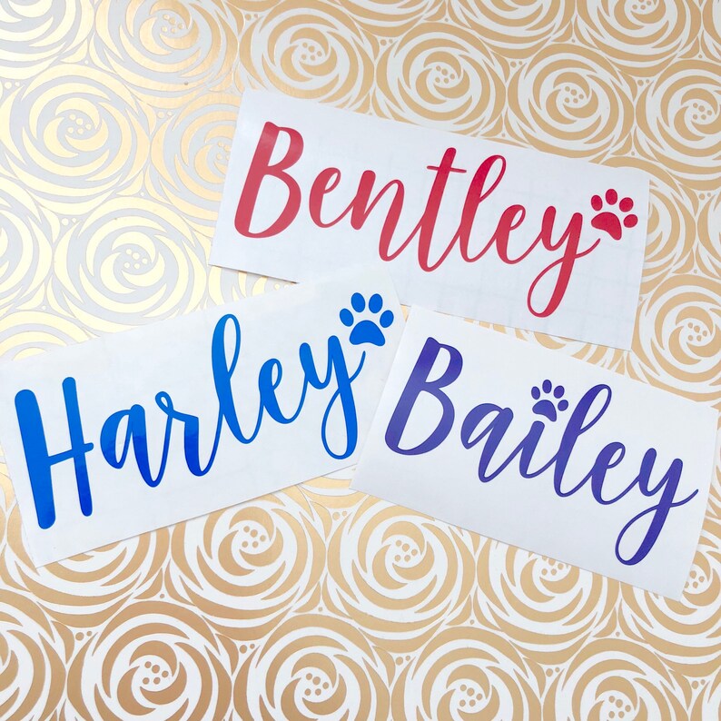 May include: Three white vinyl stickers with paw prints and names written in cursive. The names are "Bentley" in red, "Harley" in blue, and "Bailey" in purple.