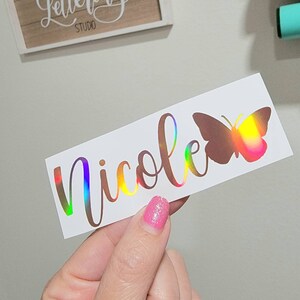 May include: A white sticker with the name "Nicole" written in a cursive font with a holographic finish. A holographic butterfly is to the right of the name.
