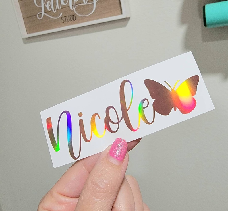 May include: A white sticker with the name "Nicole" written in a cursive font with a holographic finish. A holographic butterfly is to the right of the name.