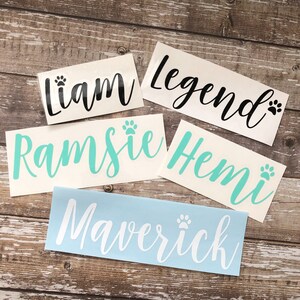 May include: Five personalized pet name decals with paw prints. The names are Liam, Legend, Ramsie, Hemi, and Maverick. The decals are on a wood background.