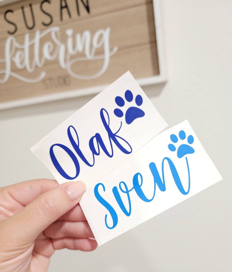 May include: Two white vinyl stickers with blue paw prints and blue cursive lettering. The stickers say "Olaf" and "Sven".