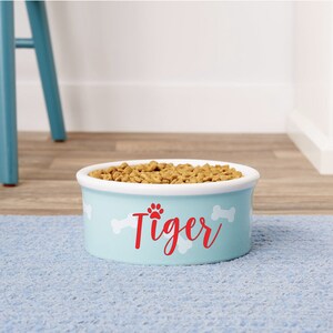 May include: A light blue ceramic dog bowl with white bone and paw print designs. The bowl has the name "Tiger" printed in red on the front.