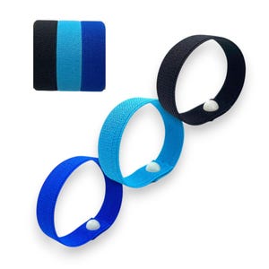 Puede incluir: Tres pulseras de tela en tonos azules y negros, cada una con un cierre de botón a presión blanco. Un pequeño cuadrado muestra las variaciones de color: negro, azul claro y azul oscuro. Las pulseras están diseñadas para la comodidad y el estilo.