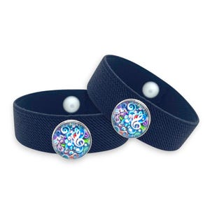 Puede incluir: Dos brazaletes elásticos azul marino con un botón de perla blanca y un diseño de nota musical colorido. El diseño de la nota musical presenta una clave de sol y otros símbolos musicales en tonos azules, morados y verdes.