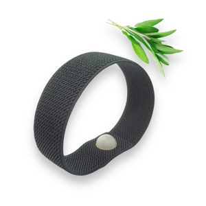 Op de afbeelding: Een donkergrijze elastische band met een witte knoopsluiting. De band is lusvormig en lijkt gemaakt van een geweven materiaal. Een takje groene salie is bij de band geplaatst.