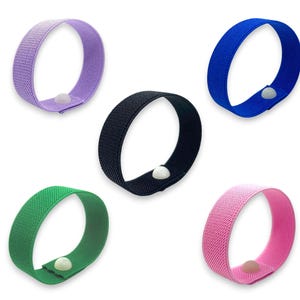 Puede incluir: Cinco bandas elásticas de colores en morado, negro, azul, verde y rosa. Cada banda tiene un cierre blanco en forma de botón. Las bandas están dispuestas sobre un fondo blanco.