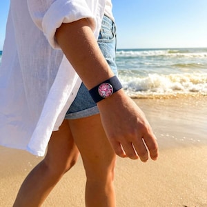 Puede incluir: Pulsera de tela azul marino con diseño floral. La pulsera se lleva en la muñeca de una persona. El diseño floral es rosa y blanco. El fondo es una playa con el océano al fondo.