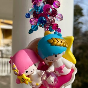 May include: A pink and blue character charm keychain with a star design. The charm features two characters, one pink and one blue, holding a clear orb. The keychain is adorned with a cluster of colorful beads.